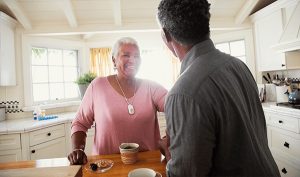 A woman wearing a Lifeline RI medical alert button is standing in her kitchen with a cup of coffee on the counter smiling a a man
