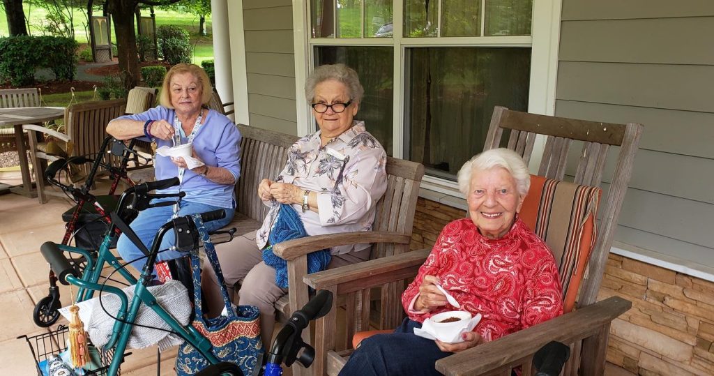 Three women sit together in comfortable chairs on the front porch of Tamarisk Senior Living