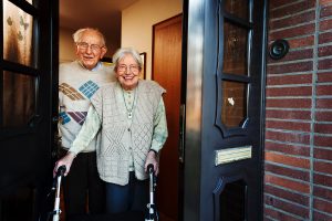 A senior couple stands in their open doorway smiling, the woman has a walker and is wearing a Lifeline RI on-the-go button